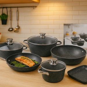 A full set of dark grey, granite-look Stovewell forged cookware displayed on a wooden kitchen counter, including casseroles, a frying pan with salmon, a wok, and a grill pan.