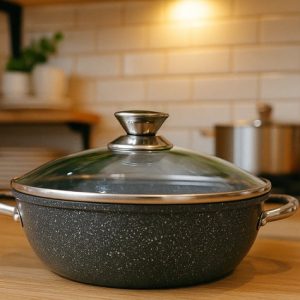 Shallow, wide dark grey non-stick casserole pot with a glass lid, stainless steel handles, and a stainless steel lid knob, sitting on a wooden counter in a kitchen.