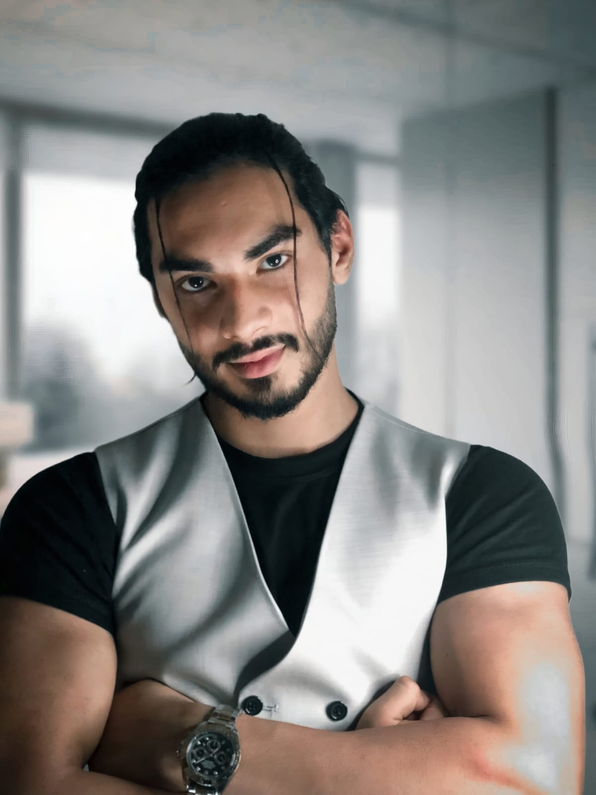 Confident Dubai-based businessman wearing a modern vest and black shirt, standing with folded arms in a corporate office setting.
